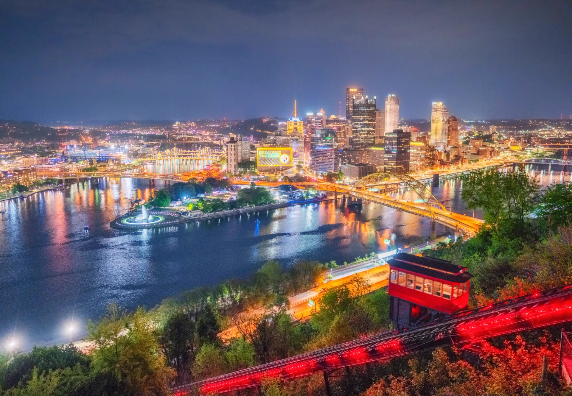 Pittsburgh skyline and Point State Park fountain at night, with a red incline car in the foreground
